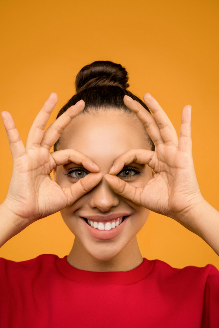 Playful portrait of a woman smiling and making hand glasses against a vibrant orange background.